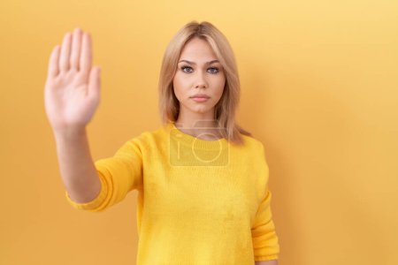 Téléchargez les photos : Jeune femme caucasienne portant un pull jaune qui arrête de chanter avec la paume de la main. expression d'avertissement avec geste négatif et sérieux sur le visage. - en image libre de droit