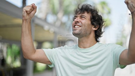 Téléchargez les photos : Jeune homme hispanique souriant confiant regardant le ciel avec un geste gagnant dans la rue - en image libre de droit