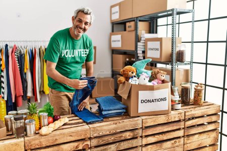 Foto de Hombre de pelo gris de mediana edad voluntario sonriendo confiado plegable jeans en el centro de caridad - Imagen libre de derechos