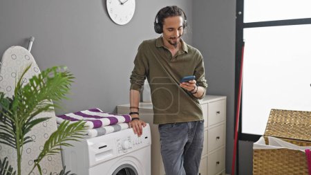 Téléchargez les photos : Jeune homme hispanique écoutant de la musique attendant la machine à laver à la buanderie - en image libre de droit