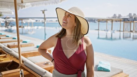 Téléchargez les photos : Une femme souriante coiffée d'un chapeau de soleil jouit d'une ambiance luxueuse au bord de la piscine dans une station tropicale surplombant la mer. - en image libre de droit