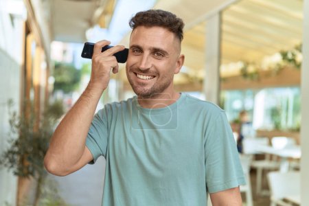 Foto de Joven hombre hispano sonriendo confiado escuchando mensaje de audio desde el teléfono inteligente en la terraza de la cafetería - Imagen libre de derechos