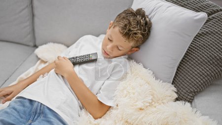 Blond boy holding tv remote control lying on sofa sleeping at home