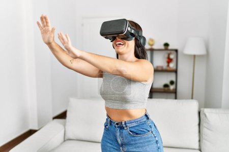 Téléchargez les photos : Jeune belle femme hispanique jouer à un jeu vidéo en utilisant des lunettes de réalité virtuelle à la maison - en image libre de droit