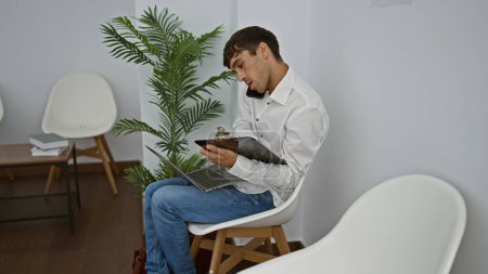 Téléchargez les photos : Portrait d'un jeune homme hispanique sérieux, un professionnel attrayant travaillant à l'intérieur. il est le patron, assis sur une chaise à l'aide d'un ordinateur portable et smartphone, gérer le succès dans une salle d'attente occupée. - en image libre de droit