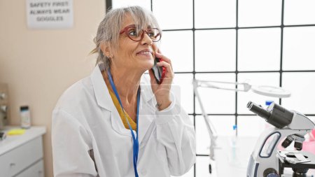 Une femme mûre en blouse de laboratoire sourit en parlant au téléphone dans un laboratoire lumineux.