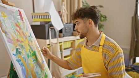 Photo for Handsome young arab man passionately immerses in his artistic journey, drawing on canvas at a bustling art studio amid fellows students. - Royalty Free Image