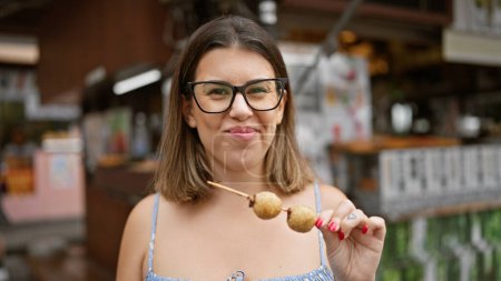 Photo for Beautiful hispanic woman in glasses relishing a traditional japanese sesame ball on a stick, immersing into the authentic street food culture of kyoto city - Royalty Free Image