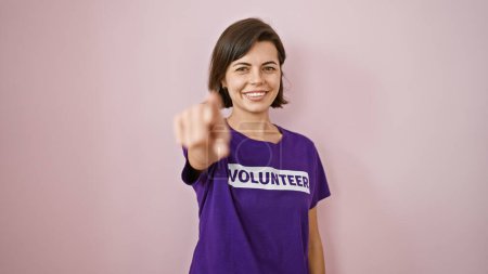 Photo for Smiling young volunteer, beautiful hispanic woman playfully pointing at you over isolated pink background, radiating a sense of unity - Royalty Free Image