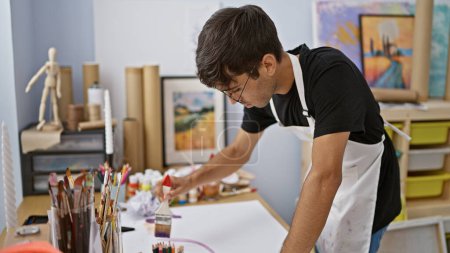 Photo for Young, handsome hispanic man seriously concentrating on drawing art in studio, brushes and palette at the ready - Royalty Free Image