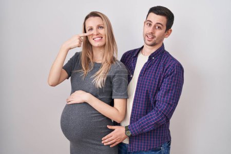 Photo for Young couple expecting a baby standing over white background pointing with hand finger to face and nose, smiling cheerful. beauty concept - Royalty Free Image