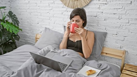 Foto de Mujer hispana hermosa joven usando el ordenador portátil hablando en el teléfono inteligente desayunando en el dormitorio - Imagen libre de derechos