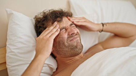 Téléchargez les photos : Jeune homme hispanique souffrant de maux de tête couché sur le lit torse nu dans la chambre - en image libre de droit