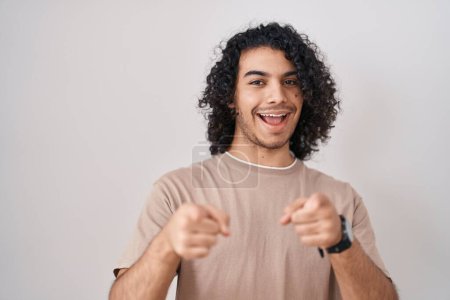 Téléchargez les photos : Homme hispanique avec les cheveux bouclés debout sur fond blanc pointant vers vous et la caméra avec les doigts, souriant positif et joyeux - en image libre de droit