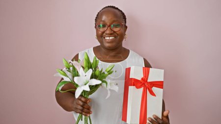 Foto de Confiada mujer afroamericana llena de alegría, sonriendo de par en par, la celebración de regalo de cumpleaños y exuberante ramo, contra el fondo aislado rosa. - Imagen libre de derechos