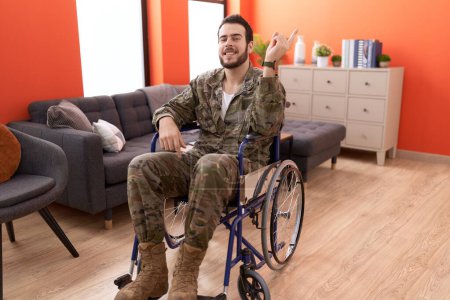 Foto de Joven soldado hispano herido sentado en silla de ruedas sonriendo feliz señalando con la mano y el dedo hacia un lado - Imagen libre de derechos