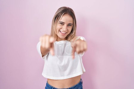 Foto de Mujer rubia joven de pie sobre el fondo rosa que apunta a usted y la cámara con los dedos, sonriendo positiva y alegre - Imagen libre de derechos
