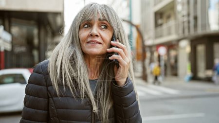 Mature hispanic woman with grey hair talking on phone on urban street.
