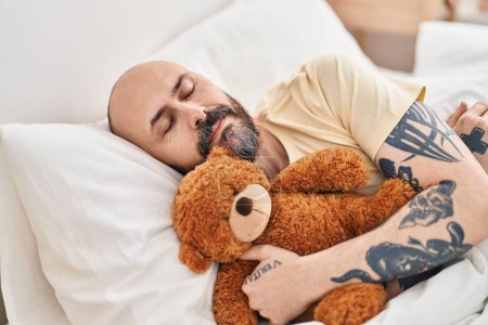 Photo for Young bald man hugging teddy bear lying on bed sleeping at bedroom - Royalty Free Image