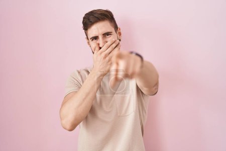 Foto de Hombre hispano con barba de pie sobre fondo rosa riéndose de ti, señalando con el dedo a la cámara con la mano sobre la boca, expresión de vergüenza - Imagen libre de derechos