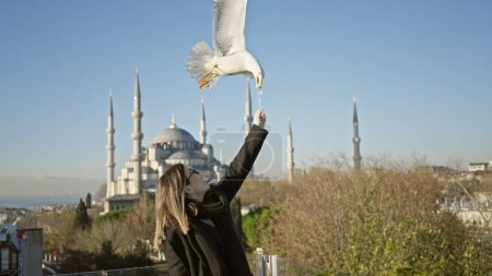 Téléchargez les photos : Une jeune femme nourrit une mouette avec une mosquée sultan ahmet en toile de fond lors d'une journée ensoleillée istanbul - en image libre de droit