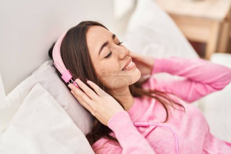 Foto de Joven mujer hispana hermosa escuchando música tumbada en la cama en el dormitorio - Imagen libre de derechos