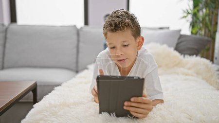Photo for Adorable blond boy, bursting with confidence, enjoying the fun of using a touchpad, is lying comfortably on the sofa at home, a picture of relaxed joy and positive lifestyle. - Royalty Free Image
