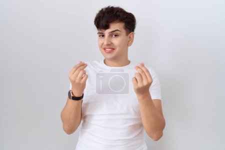 Téléchargez les photos : Jeune homme non binaire portant un t-shirt blanc décontracté faisant geste d'argent avec les mains, demandant un paiement de salaire, entreprise millionnaire - en image libre de droit