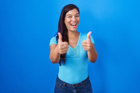 Téléchargez les photos : Jeune femme hispanique debout sur fond bleu signe de succès faisant geste positif avec la main, pouces levés souriant et heureux. expression joyeuse et geste gagnant. - en image libre de droit