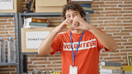 Photo for Young hispanic man activist wearing volunteer uniform doing heart gesture at charity center - Royalty Free Image