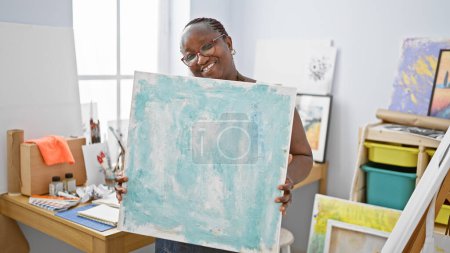 Téléchargez les photos : Artiste afro-américaine confiante avec des tresses et des lunettes, souriant joyeusement et tenant toile, prête à dessiner dans un atelier d'art confortable. - en image libre de droit