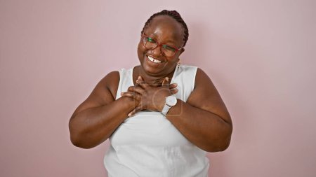 Foto de Alegre mujer afroamericana de tamaño grande con trenzas, radiante con confianza, las manos sobre el pecho, de pie sobre un hermoso fondo aislado rosa - Imagen libre de derechos