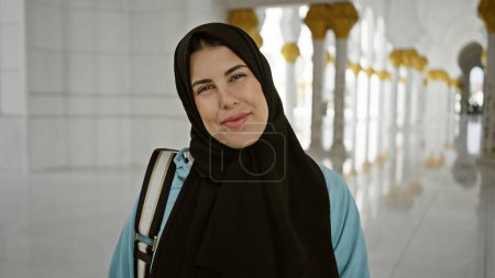 Photo for A beautiful young adult woman in a hijab smiles inside the ornate architecture of a mosque in abu dhabi. - Royalty Free Image