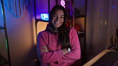 Photo for Smiling young woman with crossed arms wearing headphones in a neon-lit gaming room at night - Royalty Free Image