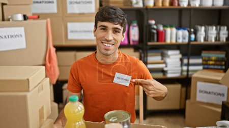 Photo for Confident hispanic young man, sitting at a table in the heart of a bustling charity center, cheerfully pointing to his volunteer badge with a big, infectious smile - Royalty Free Image