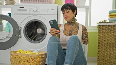 Téléchargez les photos : Femme hispanique avec bras amputé utilisant smartphone assis par machine à laver à la buanderie - en image libre de droit