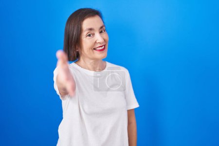 Foto de Middle age hispanic woman standing over blue background smiling friendly offering handshake as greeting and welcoming. successful business. - Imagen libre de derechos