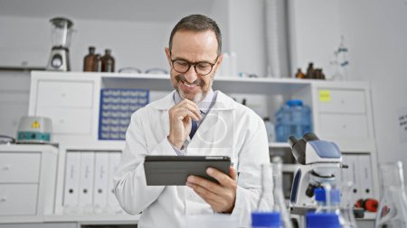 Foto de Alegre hombre de mediana edad, un científico de pelo gris, alegremente usando un touchpad mientras conduce su investigación en la vibrante atmósfera de su laboratorio. - Imagen libre de derechos