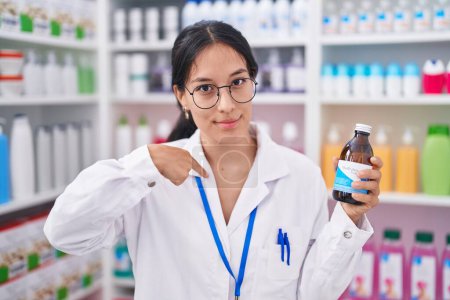 Téléchargez les photos : Jeune femme hispanique travaillant à la pharmacie pharmacie tenant sirop pointant du doigt un sourire heureux et fier - en image libre de droit