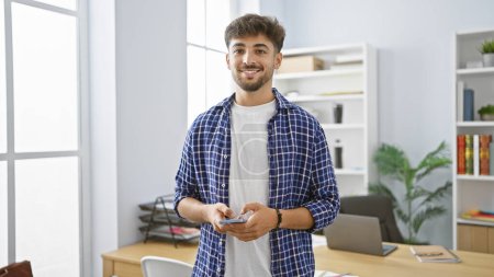 Foto de Hombre árabe joven confiado, radiante con una expresión feliz, absorto en el trabajo, gestiona sin esfuerzo las tareas de negocio en su teléfono inteligente, de pie en una elegante oficina en el interior. - Imagen libre de derechos