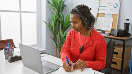 Photo for An african american woman works diligently in a modern office, wearing a headset and taking notes. - Royalty Free Image