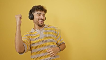 Téléchargez les photos : Cool, confiant jeune homme arabe grooving à la musique sur son casque. dansant heureux seul devant un mur jaune isolé, rayonnant de positivité et de bonheur. - en image libre de droit