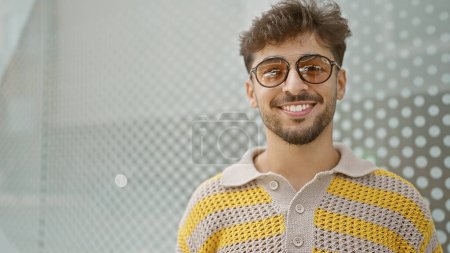 Photo for Confident young arab man, radiating joy and positivity, is standing outdoors on a city street, resplendently handsome in his sunglasses, his contagious smile lighting up the urban background. - Royalty Free Image
