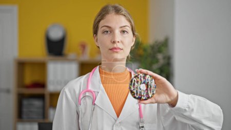 Téléchargez les photos : Jeune femme blonde diététicienne tenant un beignet avec un visage sérieux à la clinique - en image libre de droit