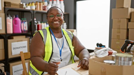 Téléchargez les photos : Femme afro-américaine souriante avec des tresses, une bénévole dévouée, assise à la table du centre de bienfaisance, absorbée par la prise de notes - en image libre de droit