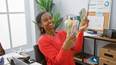Téléchargez les photos : Femme africaine souriante examinant les rands sud-africains dans un cadre de bureau moderne - en image libre de droit
