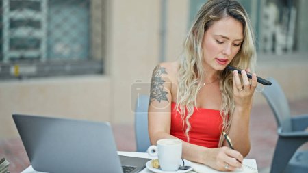 Photo for Young blonde woman sending voice message by smartphone taking notes at coffee shop terrace - Royalty Free Image
