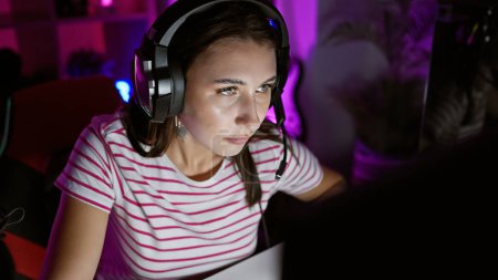 Foto de Mujer joven enfocada con auriculares en una sala de juegos oscura iluminada por luces de colores - Imagen libre de derechos