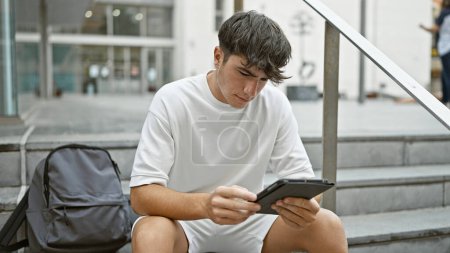 Photo for Cool young hispanic teenager, a serious student, casually sitting on university stairs, engrossed in his laptop screen - concentrated on touchpad, embodying smart tech lifestyle outdoors. - Royalty Free Image