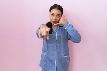 Téléchargez les photos : Jeune femme arabe en pyjama bleu souriant faisant parler au téléphone geste et vous pointant du doigt. Appelle-moi. - en image libre de droit